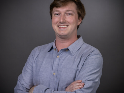 Headshot of Paul Mayo wearing a blue shirt in front of a black background. His arms are crossed and he is smiling. 