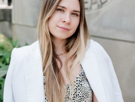 Angelina Bilokon standing outdoors in front of a university building, wearing a patterned top and a white blazer draped over her shoulders. She faces the camera with a slight smile.