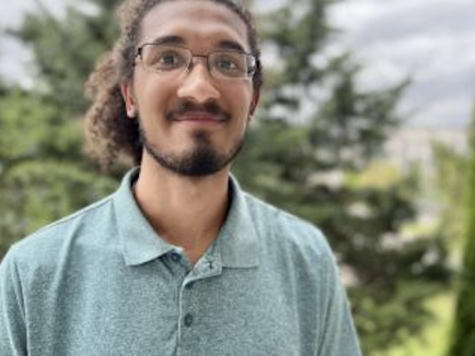 Jason Putnam stands outdoors in front of blurred trees and a cloudy sky, wearing a light teal polo shirt and smiling at the camera.