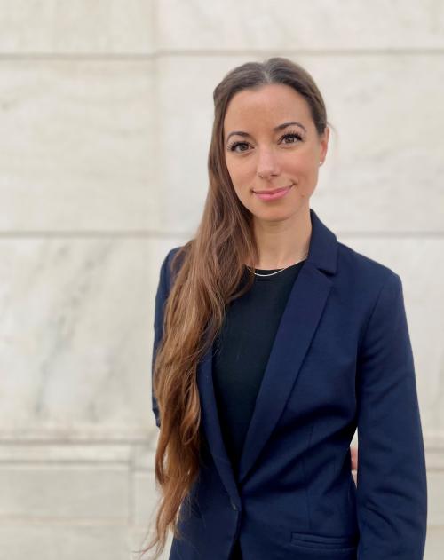 A woman with long brown hair and a blue blazer looks at the camera