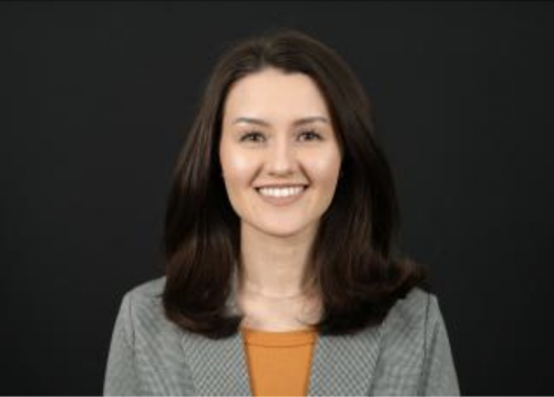 Charlier Fisher in front of a black background with an orange shirt and gray blazer.