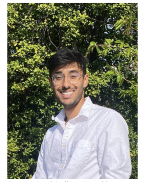 A young man with short dark hair and glasses smiles at the camera while standing outdoors in front of leafy green trees. He is wearing a white button-down shirt and is lit by natural sunlight.