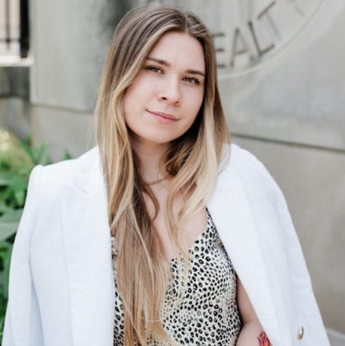 Angelina Bilokon standing outdoors in front of a university building, wearing a patterned top and a white blazer draped over her shoulders. She faces the camera with a slight smile.
