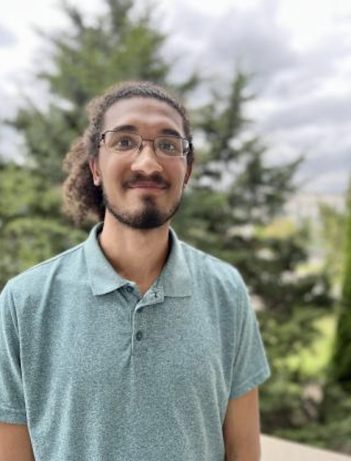 Jason Putnam stands outdoors in front of blurred trees and a cloudy sky, wearing a light teal polo shirt and smiling at the camera.