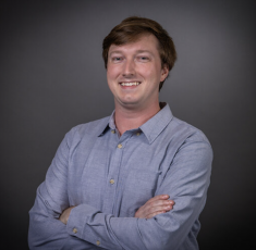 Headshot of Paul Mayo wearing a blue shirt in front of a black background. His arms are crossed and he is smiling. 