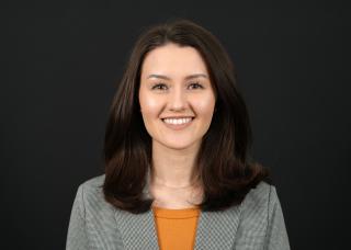 Headshot displaying a woman smiling. Woman has medium-length, dark brown hair and is wearing a gray blazer over an orange shirt. Image is cropped to display appearance from head to shoulders. 