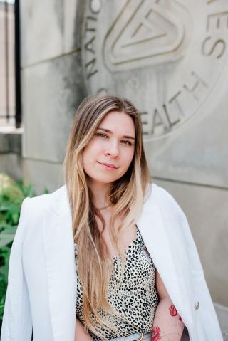 A headshot of a woman sitting outdoors in front of a stone wall with the engraved words "National Institutes of Health."